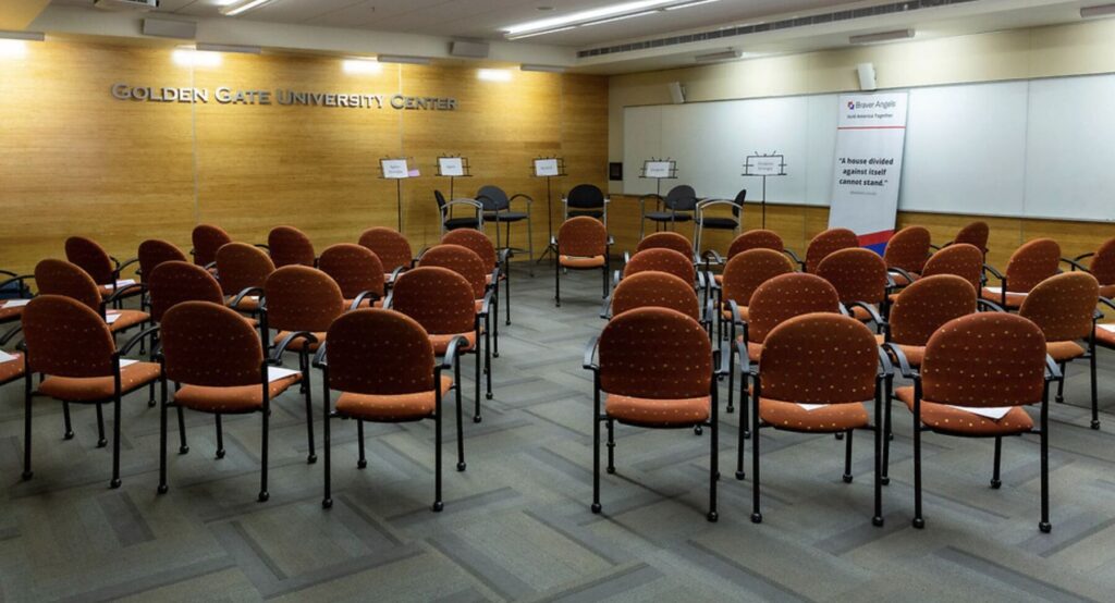 A room is filled with empty chairs arranged for an audience; in front of the room are five chairs with signs that say Agree Strongly, Agree, Neutral, Disagree, Disagree Strongly.