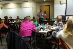 A man in a plaid shirt speaks to an audience of people seated at tables in a university conference room.